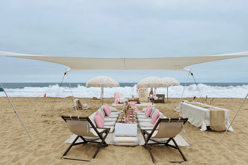 Neutral beach baby shower setup featuring low picnic seating with blush pillows, boho umbrellas, pampas decor, and ocean backdrop in Newport Beach.